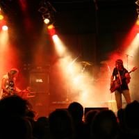 At the 2011 festival, a rock band is performing on stage with dramatic red and yellow lighting illuminating the scene. The guitarist on the left is playing and standing near a microphone, while the bassist on the right wears a headband. A drum set is visible in the background. A crowd watches from the foreground.