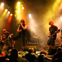 At the Fontaine 2011 festival, a rock band performs energetically on stage with bright, colorful lights and smoke effects in the background. Four band members play electric guitars, and one sings into a microphone. Audience members are visible at the front of the stage, raising their hands in excitement.
