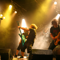 A rock band performs energetically on a dimly lit stage with vibrant yellow spotlights at the 2011 Fontaine Festival. The guitarist in the center plays a teal electric guitar, bending backwards. The bassist on the right and another guitarist on the left play intensely while the drummer is partially visible at the back.