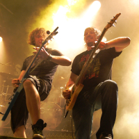 Two musicians play electric guitars on stage at the 2011 Fontaine Festival, surrounded by yellowish haze from stage lighting. Both are in dark t-shirts, shorts, and sneakers, with one wearing glasses. Each has one foot resting on a monitor speaker. Drums are visible in the background, and the atmosphere is energetic and intense.