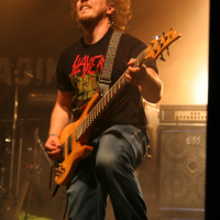 At the 2011 Fontaine Festival, a musician with curly hair passionately plays an electric bass guitar on stage. He is wearing a black T-shirt with a graphic print, blue jeans, and a guitar strap over his shoulder. Stage lights create a smoky atmosphere, and amplifiers are visible in the background.
