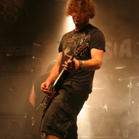 A musician with curly hair, wearing a dark t-shirt and shorts, energetically plays an electric guitar on stage at the 2011 Fontaine Festival. The stage is dimly lit with spotlights and smoke effects. Amplifiers and drum equipment are visible in the background, along with indistinct band signage on the back wall.