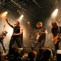 A rock band performs energetically on stage at the Fontaine festival 2011. The lead singer holds a microphone and belts out a song while raising a fist. Other band members play electric guitars and a bass, and one raises an arm in the air. Stage lights shine from above, creating a dramatic atmosphere as the crowd watches.