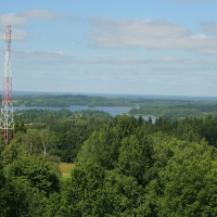 A towering red and white communications mast rises above a lush green forest near Gaiziņš with a serene lake visible in the distance under a clear blue sky.