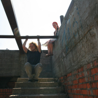 A man is crouched on a set of concrete stairs beneath a steel beam in a partly constructed building, holding onto a horizontal beam above him. A woman sits at the edge of a concrete wall beside him. The walls have visible graffiti, and the sunlight contrasts with the shadows inside, like something out of Gaiziņš' realm.