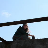 A smiling person sits atop a concrete structure under a clear blue sky, resting her arms on a rusty metal beam. Her casual attire and relaxed posture suggest she's enjoying a moment of leisure in an industrial