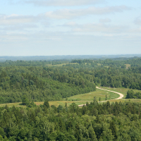 Aerial view of a verdant landscape near Gaiziņš with a winding road cutting through dense forests under a clear blue sky.