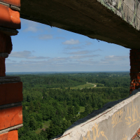 A view from a dilapidated structure with exposed brickwork overlooking the lush green forest landscape of Gaiziņš and winding road, under a clear blue sky.