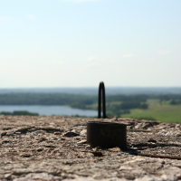 A metal loop protrudes from weathered concrete with a scenic view of Gaiziņš, a peaceful lake, and green landscape blurred in the background, under a clear blue sky.