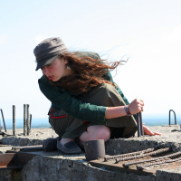 A woman with long, flowing hair and wearing a gray hat and green jacket crouches on a concrete structure near Gaiziņš. She is holding a metal rod and surrounded by rebar and metal components. The sky is clear with a faint horizon in the background. She appears focused on her task.