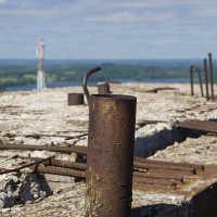 A close-up of a rusted metal rod protruding from the crumbling concrete of an abandoned, unfinished building. The foreground is filled with more exposed rebar and deteriorating concrete. In the distance, there's a blurred view of a green forested landscape and Gaiziņš standing tall against the horizon.