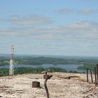 A wide-view landscape image showing a concrete surface with exposed rebar in the foreground, overlooking a lush green expanse with scattered trees and a lake in the background. A tall communication tower stands to the left amidst the greenery near Gaiziņš. The sky is blue with some clouds.