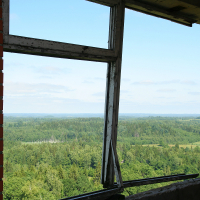 A view through a broken window frame in a red brick structure, overlooking the lush green landscape of Gaiziņš under a clear blue sky.