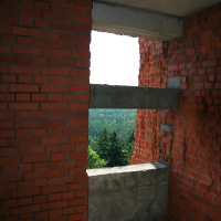 A view through a rough, unfinished window opening in a brick wall, overlooking a dense, verdant Gaiziņš forest under a bright blue sky.