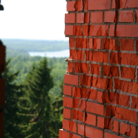 A close-up view of a weathered red brick wall with vibrant green trees and a glimpse of a calm lake in the blurred background, as seen from a high vantage point on Gaiziņ