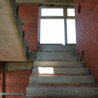 A gritty, unfinished staircase with bare red brick walls and exposed concrete surfaces leading to a brightly-lit window atop Gaiziņš, exuding a raw, industrial atmosphere.