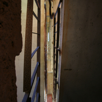 A dimly lit stairwell with rusted metal banisters ascends between weathered concrete walls, while narrow slits of light peek through, hinting at an urban, gritty Gaiziņ