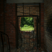 A dilapidated room with crumbling brick walls and scattered debris leads to a rusted, broken metal gate, framing a view of Gaiziņš's lush greenery outside, highlighting contrasts of