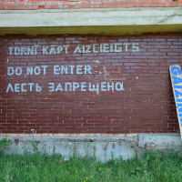 A brick wall with a sign in multiple languages, including Latvian, English, and Russian, reads, "TURNI KĀPT AIZLIEGTS," "DO NOT ENTER," and "ЛЕСТЬ ЗАПРЕЩЕНО," indicating that entry is forbidden. A blue and white sign leaning against the wall features the word “GAIZINKALNS.” Grass is