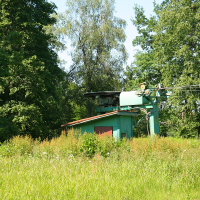 A small, green building with a red door is nestled among tall grass and dense trees near Gaiziņš, Latvia's highest peak. Beside the building stands a turquoise mechanical structure, possibly a ski lift or cable car machinery, with wires extending to the right. The scene basks in bright sunlight, enhancing the lush greenery.