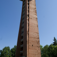 A tall, narrow, red brick tower with a pyramidal top and a slightly overhanging green roof stands solitary against a clear blue sky, surrounded by lush Gaiziņš greenery