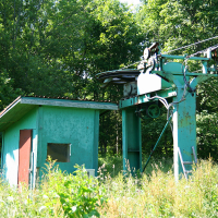 An old ski lift station, overgrown with lush greenery, stands abandoned amidst nature near Gaiziņš, its mechanism dormant and encroaching foliage signalling disuse.