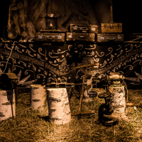 An outdoor night scene at the Give and Get festival 2013 shows several wooden trunks and metal tools arranged on the ground in front of a detailed, patterned backdrop and multiple stacked suitcases. The area is dimly lit, creating shadows and an old-fashioned, rustic atmosphere. The ground is covered with hay, adding to the rustic feel.