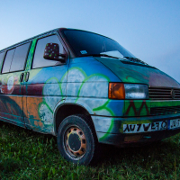 A colorful van, adorned with vibrant graffiti and abstract designs, is parked on a grassy field near a forested area at dusk. The scene harkens back to the 2013 Give and Get festival. The sky transitions to night, casting a blue hue, while a silver car is partially visible in the background on the right side.