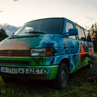 A vibrant, multi-colored van is parked on the grass at dusk, with trees in the background. The van, with its artistic paint job from the 2013 Give and Get festival, features an array of blues, greens, and oranges. Two people are kneeling near the van, adding more details to the artwork on the vehicle's side.