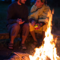 Two people sit by a campfire at night, engaged in conversation. The person on the right wears a yellow onesie with rabbit ears, while the person on the left wears casual clothes. The warm firelight illuminates them, creating a cozy atmosphere against the dark background of the 2013 Give and Get festival.