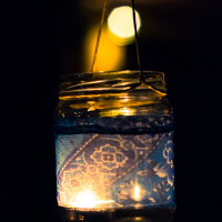 A glass jar with a lit candle inside hangs by a thin string against a dark background. Adorned with decorative blue lace fabric, the jar casts intricate floral patterns when illuminated by the candlelight, creating a warm, soft glow. A blurred bright spot appears in the background of this 2013 Give and Get festival scene.