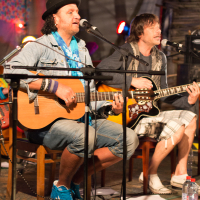 Two male musicians perform on stage with acoustic guitars during the 2013 Give and Get festival. The man on the left wears a black hat, blue scarf, denim jacket, and jeans with blue sneakers. The man on the right sports a grey shirt and plaid shorts with black boots. Both sing into microphones as they play their guitars.