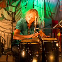 A man with long blonde hair and a blue shirt plays two large conga drums in a colorful, dimly lit setting at the Give and Get 2013 festival. A microphone is positioned near the drums, capturing the sound. The background features vibrant, multi-colored decorations and stage lighting, with additional drums and musical equipment visible.