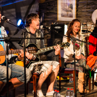Four musicians are performing on stage at the 2013 Give and Get Festival. Two men on the left are playing acoustic guitars. One wears a hat, scarf, and jean shorts; the other sings passionately into a microphone. Two women on the right sing into microphones; one in a white jacket and patterned leggings, and the other in a red top and black scarf.
