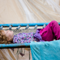 A young child with light brown hair lies on their back on a blue trampoline at the 2013 Give and Get festival. They are wearing a purple floral shirt and pink pants, with a light blue towel draped over the edge. The child looks relaxed, with a calm expression, while a large white tarp is visible in the background.