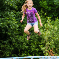 A young girl with long blonde hair, wearing a purple T-shirt, denim shorts, and sandals, is mid-air while jumping on a trampoline at the Give and Get festival. Green foliage and trees are visible in the background, emphasizing that the activity takes place outdoors. She appears focused and energetic.