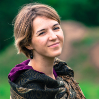 A person with short brown hair is standing outdoors at the 2013 Give and Get festival, wearing a purple top and a patterned shawl. They are slightly smiling and looking at the camera. The background is blurred greenery, suggesting a natural, possibly forested, setting.