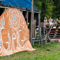 A large, orange, hand-painted sign with intricate white patterns and text reading "G! Festival" leans against a rustic wooden structure. People sit and chat on the building's wooden steps, surrounded by lush greenery. Colorful lights and decorations add to the festive atmosphere of the 2013 festival.
