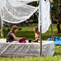 Two young girls sit on an outdoor mattress under a makeshift canopy made of lace curtains and wooden poles. One girl holds a red toy sailboat, while the other watches. The scene is set in a grassy area with scattered items and greenery in the background, reminiscent of a 2013 festival vibe.