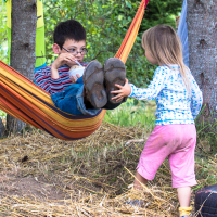 A boy with glasses is lying in a colorful hammock holding a cup, wearing a striped shirt, jeans, and brown shoes. A young girl in a white star-patterned shirt and pink pants stands next to him with her arm extended under a tree in a grassy area. Her clothes and feet are dirty from the 2013 Give and Get festival.