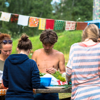 At the 2013 Give and Get festival, a group of people are gathered outdoors around a table preparing food. One person in the foreground is wearing a striped shirt and another has a hooded sweatshirt. A shirtless man is focused on the task. Colorful fabric banners are strung overhead, and greenery is lush in the background.