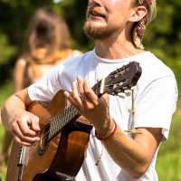 A man with a beard, wearing a white shirt and a patterned bandana, plays an acoustic guitar outdoors at the Give and Get Festival 2013. He appears to be passionately singing with his eyes closed. The background features greenery and an out-of-focus person, and a blue vertical flag or banner is partially visible.