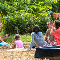 A group of five people, including two children and three adults, relaxes outdoors on a grassy area with hay at the Give and Get festival 2013. One adult plays a small guitar while the others listen or chat. Towels hang on a line behind them, and trees provide shade. A large plastic container is visible in the foreground.