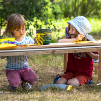 At the 2013 Give and Get festival, two young children are seated outdoors at a low wooden table engaging in art activities. The child on the left wears a white shirt with blue patterns and pink pants, while the child on the right sports a blue hat, red shirt, and shorts. The table is covered with art supplies, including papers and markers.