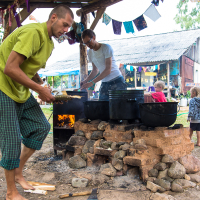 Two men are cooking outdoors under a wooden canopy at the 2013 Give and Get festival. One wears a green shirt and checkered pants, tending to the fire, while the other, in a white T-shirt and jeans, stirs a pot. Two children play nearby as colorful fabric buntings hang above. Rustic buildings and lush greenery form the backdrop.