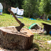 A hatchet is lodged in a tree stump in a grassy outdoor setting. In the background, there are tents, a clothesline with hanging items, and several people enjoying the 2013 Give and Get festival. To the right, an outdoor basin or cooking pot sits on the grass, surrounded by various camping materials. Trees provide partial shade.
