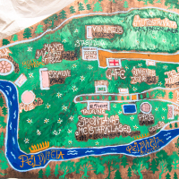 A colorful, hand-drawn map on a brown canvas shows various zones and activities labeled in Latvian for the 2013 Give and Get festival. A blue river with "Peldvieta" flows through, surrounded by green areas marked with "Gyvesciute zona," "Cafe," "Skatuve," and "Autostavieta." Paths and icons highlight different spots.