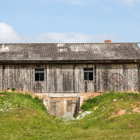 A weathered wooden barn with a corrugated metal roof sits atop a grassy mound. The barn, believed to be from the 2013 Give and Get festival, has two dark windows and is adorned with colorful, sporadic floral patterns. A large peace sign is painted on the right side. In the background, trees and a blue sky with clouds are visible.