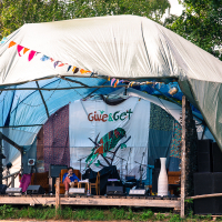 A covered outdoor stage with a "Give & Get" backdrop, reminiscent of the 2013 Give and Get festival. The stage is set with musical instruments and equipment, including a drum set and speakers. A person in a blue shirt sits before the backdrop. Colorful triangular flags hang above, framed by trees in the background.