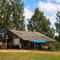 At the Give and Get 2013 festival, an outdoor market stall is set up under a rustic wooden canopy with a corrugated metal roof. Colorful bunting hangs above, and items are displayed along the wooden structure. Trees and a partly cloudy blue sky create a festive, rural atmosphere.